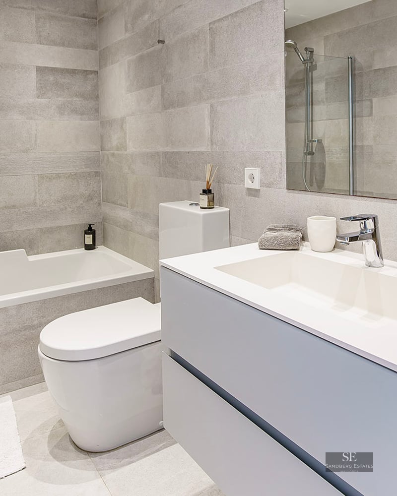 Contemporary bathroom featuring grey stone-effect tiles, a white bathtub, toilet, and modern vanity with a large mirror.