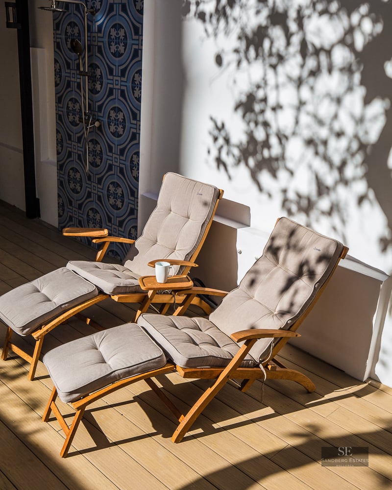 Two wooden lounge chairs on a deck next to a decorative blue tiled outdoor shower with leaf shadows on the wall.