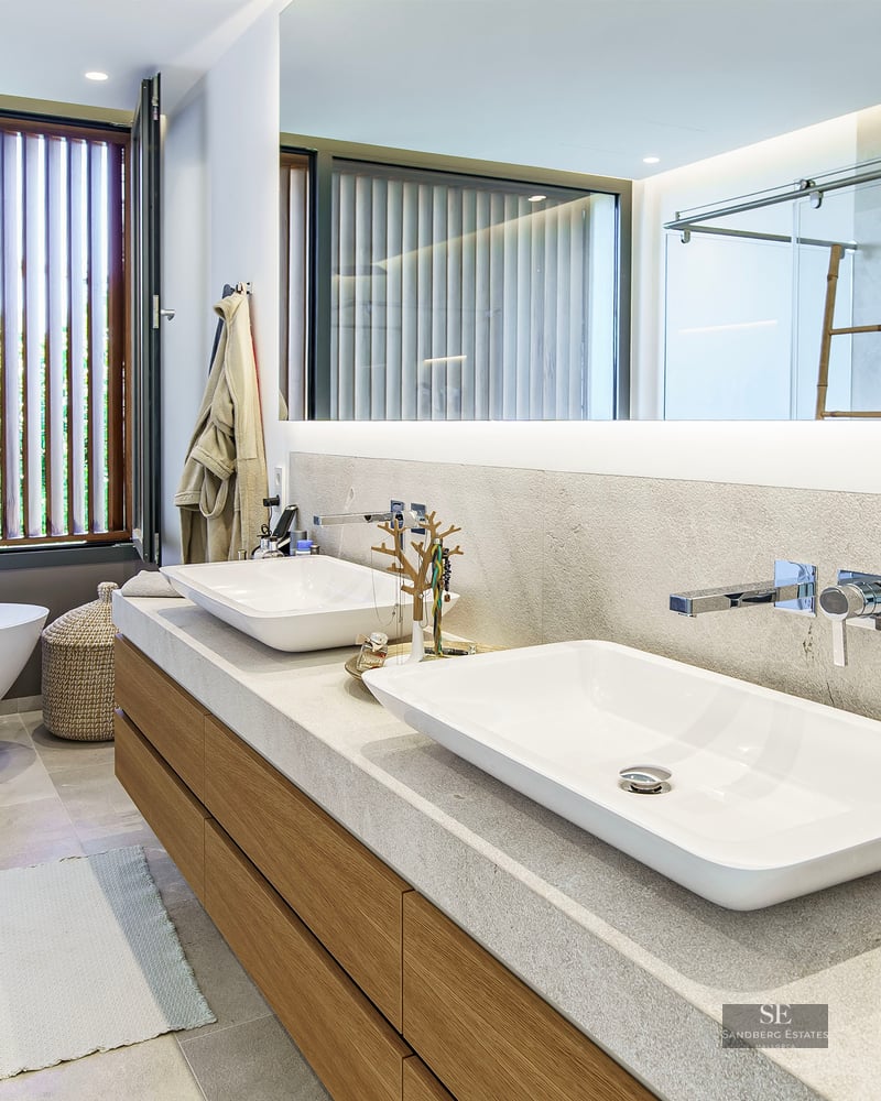 Salle de bain moderne avec double vasque, grand miroir éclairé et baignoire autoportante près d'une fenêtre à persiennes.