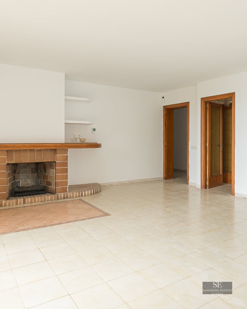 Empty living room with beige floor tiles, white walls, a brick fireplace, and a kitchen pass-through window.