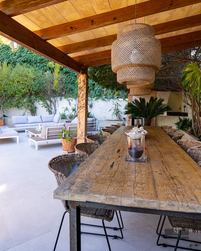 Long wooden dining table under a pergola with wicker chairs and a pendant light next to an outdoor lounge.