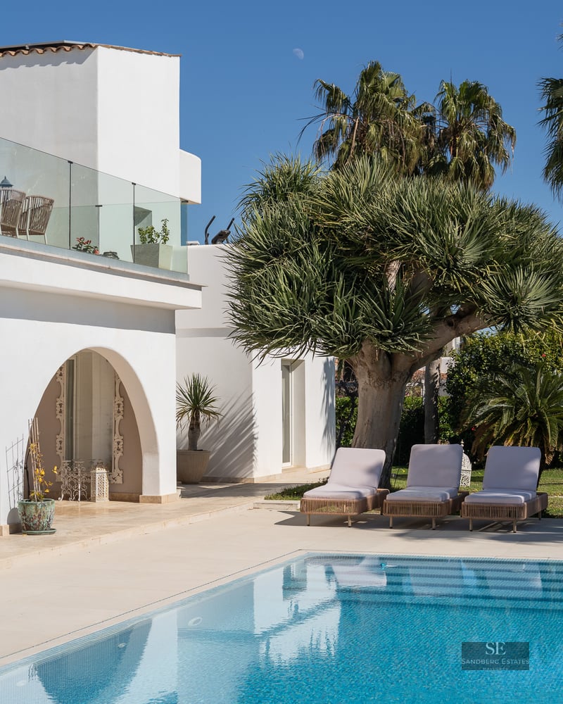 A bright swimming pool next to a white villa with three sun loungers and tropical palm trees under a blue sky.