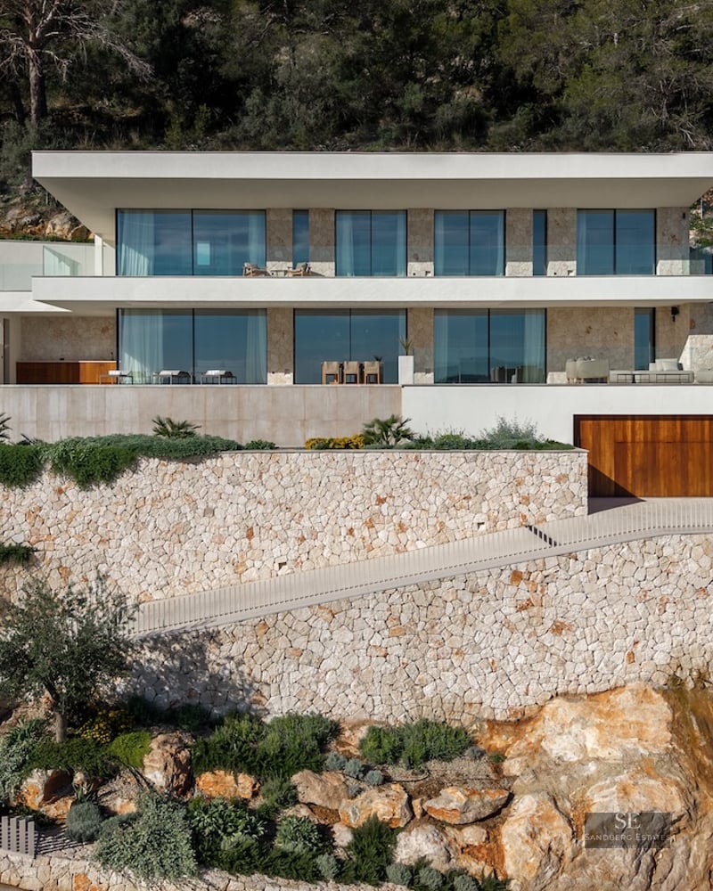 Modern white villa built into a rocky hillside with dry stone walls, large floor-to-ceiling windows, and a curved driveway.