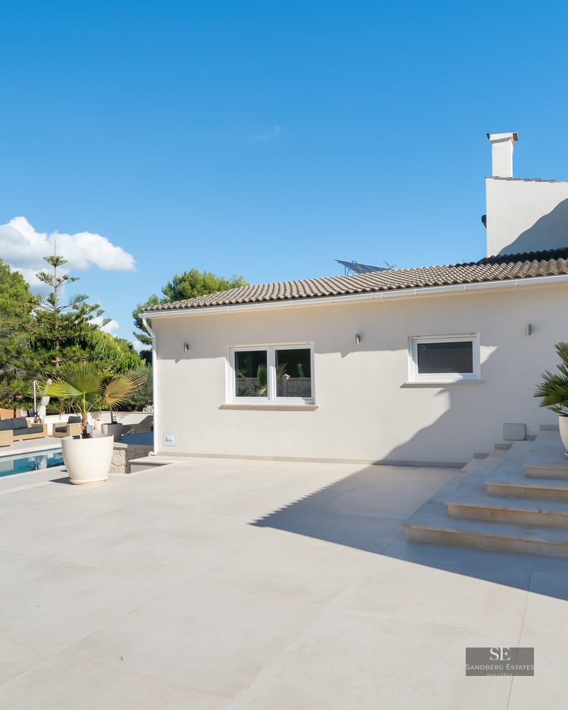 Villa blanche moderne avec piscine, grande terrasse en pierre et palmiers en pots sous un ciel bleu clair.