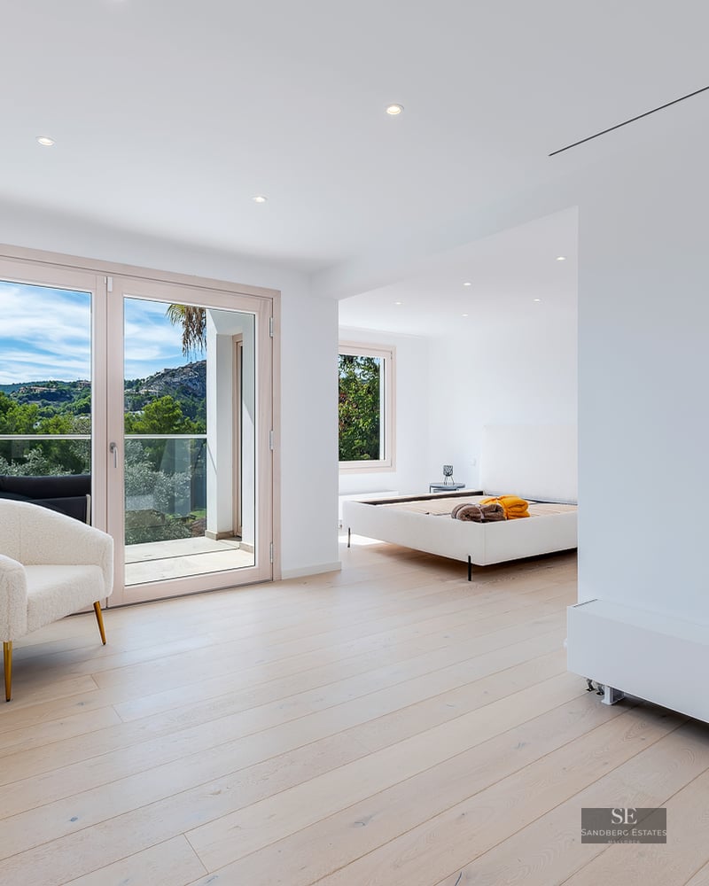 Bright master bedroom with light wood floors, a designer armchair, balcony access, and an ensuite bathroom visible.