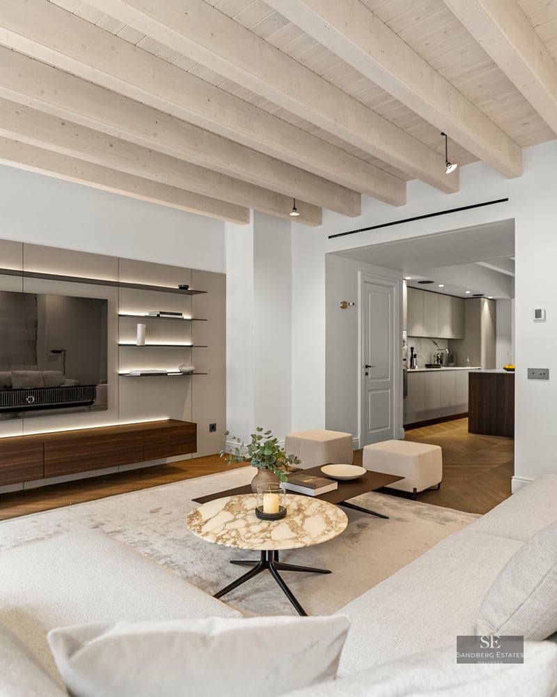 Modern living room with white sofas, marble coffee table, wood-beamed ceiling, and built-in TV wall.