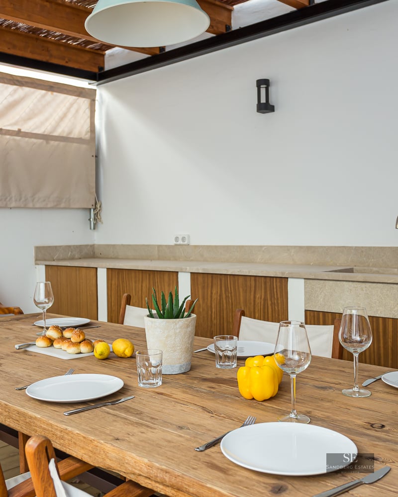 Long wooden table set for several guests on a covered terrace with an outdoor kitchen in the background.
