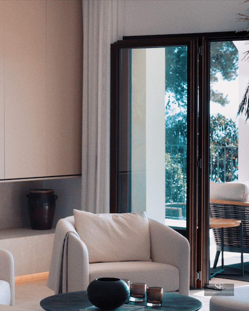 Bright minimalist living room with white armchairs, integrated TV, and large glass doors opening to a sunny terrace.