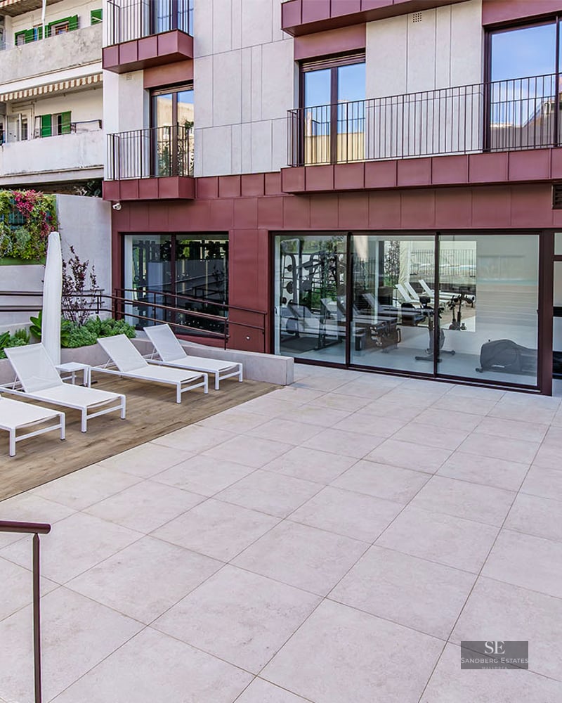 Modern outdoor terrace with white sun loungers, a vertical garden wall, and a view into a glass-walled gym.