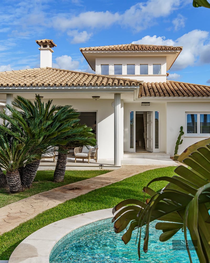 White Mediterranean villa with a terracotta roof, a lush green lawn, palm trees, and a turquoise swimming pool.