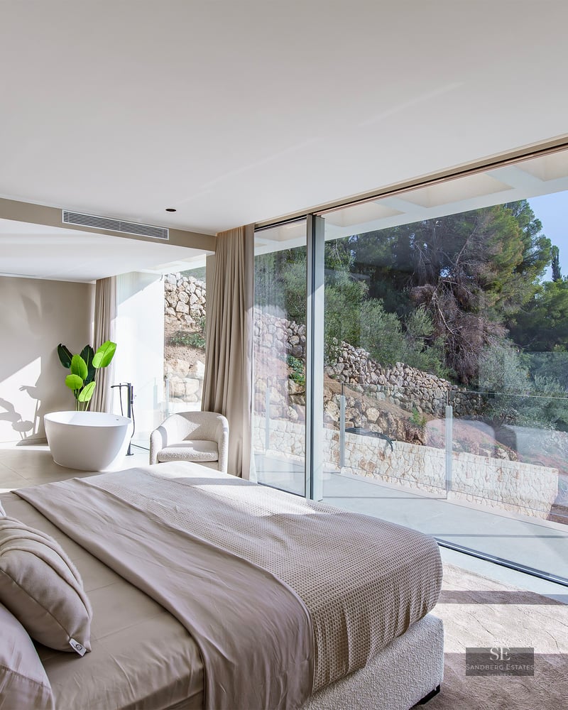 Modern bedroom with floor-to-ceiling windows, a large bed in neutral tones, and an ensuite bathtub with garden views.