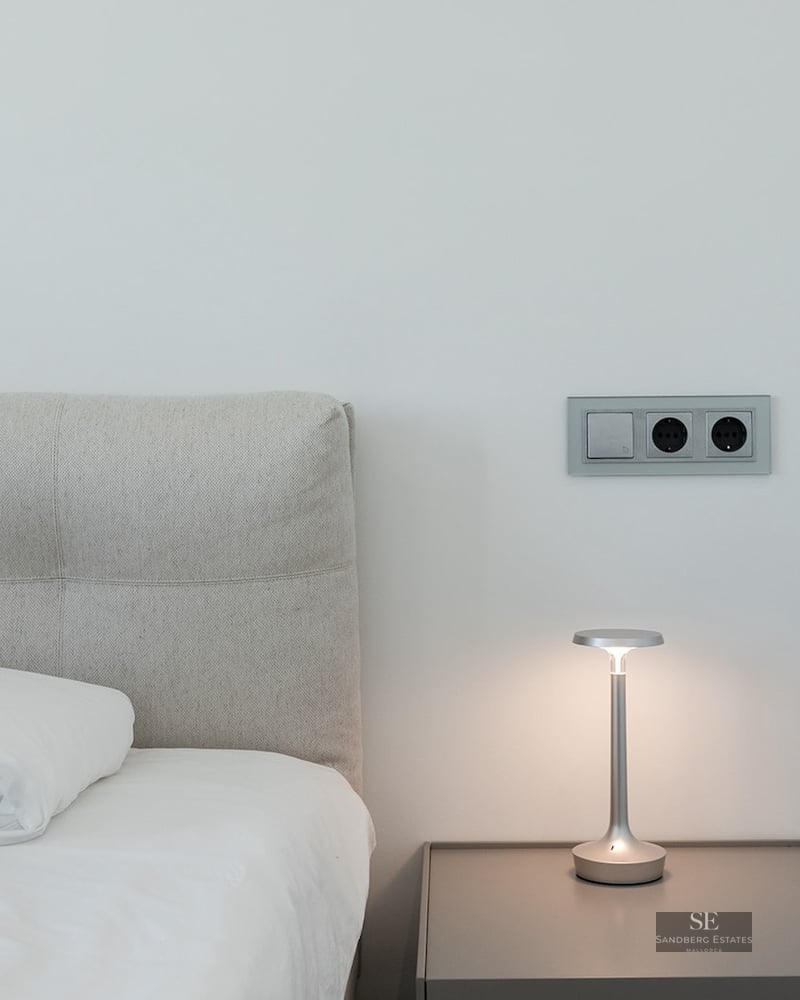 Close-up of a bed with grey headboard, nightstand with a designer lamp, and modern electrical sockets on a white wall.