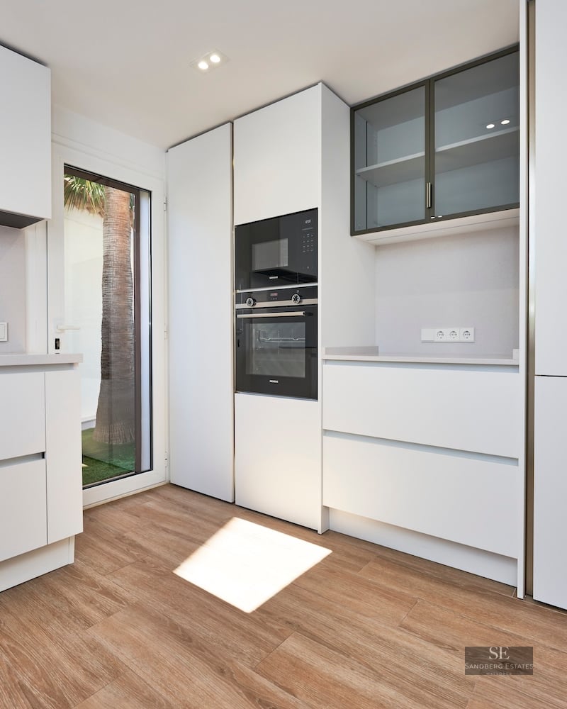 Minimalist white kitchen with integrated appliances, wood-look floors, and a glass door showing a palm tree.