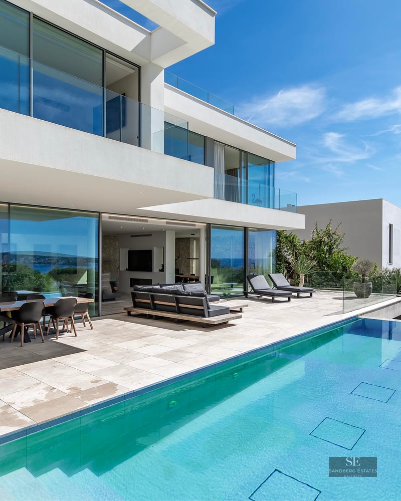 Modern white villa with an outdoor swimming pool, terrace furniture, and a panoramic view of the sea under a clear blue sky.
