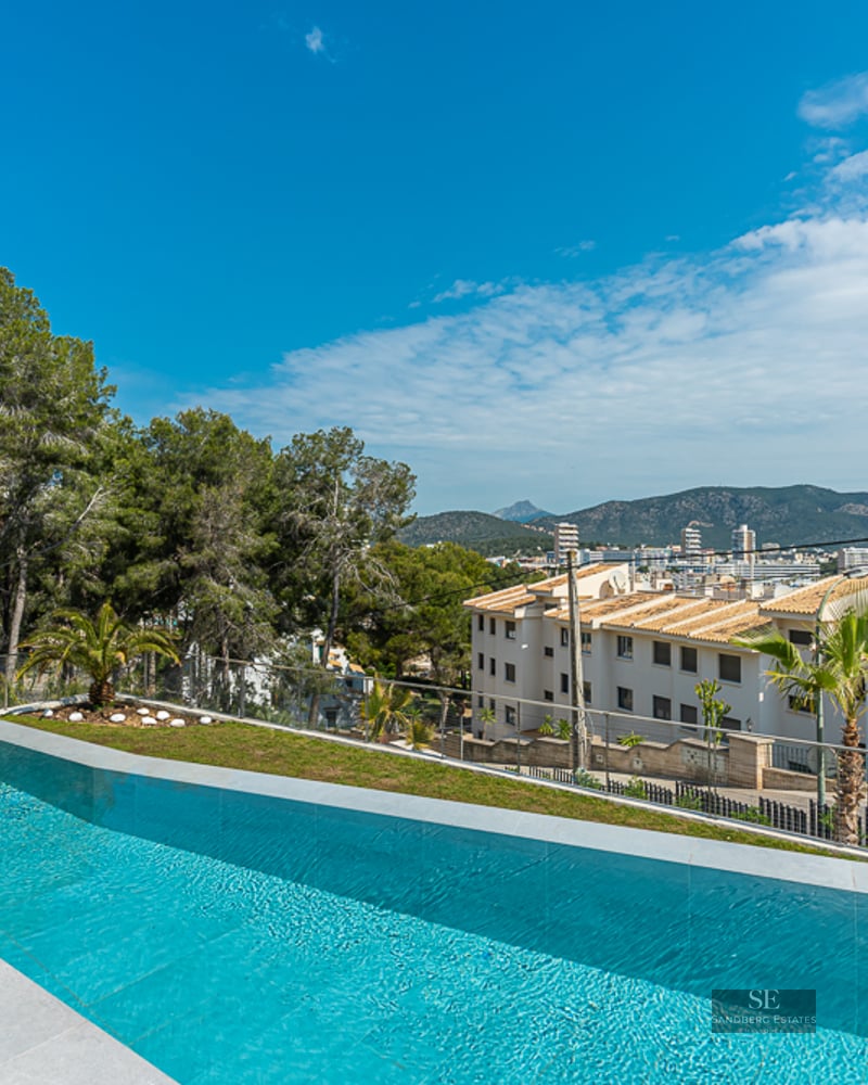 Luxury infinity swimming pool with stone tiling, overlooking a coastal town, mountains, and the sea under a clear blue sky.