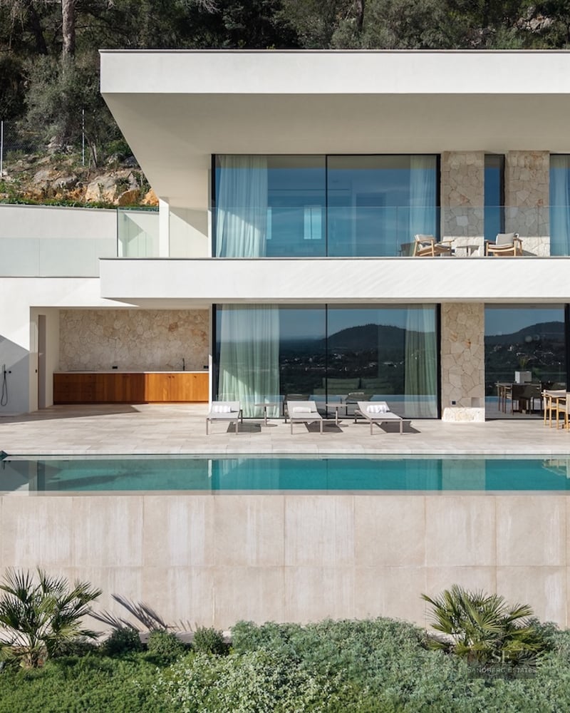 Modern two-story villa with a turquoise infinity pool, stone walls, large glass windows, and a sunny terrace.