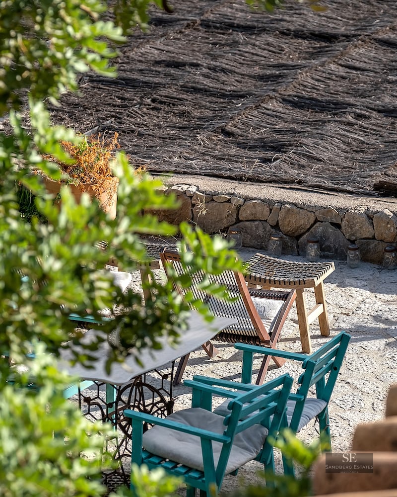 Outdoor stone terrace with teal wooden chairs, vintage table, and a thatched roof seen through greenery.