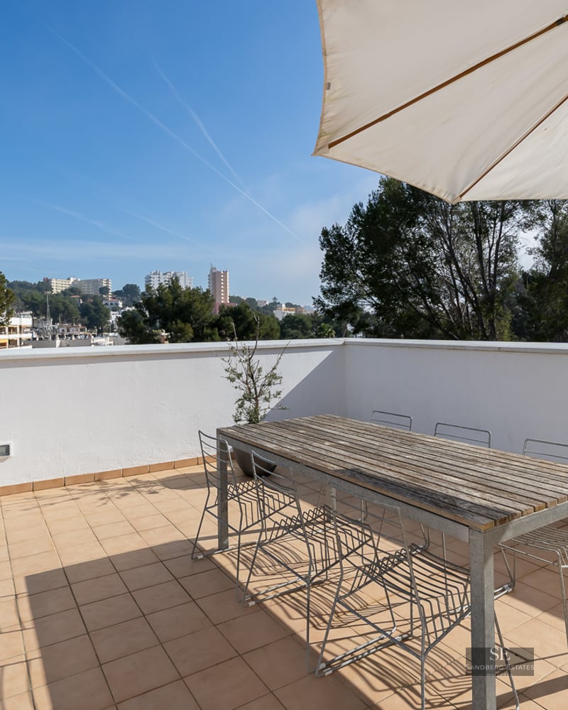Spacious rooftop terrace with a large wooden table, wire chairs, and a white sun umbrella overlooking a city.
