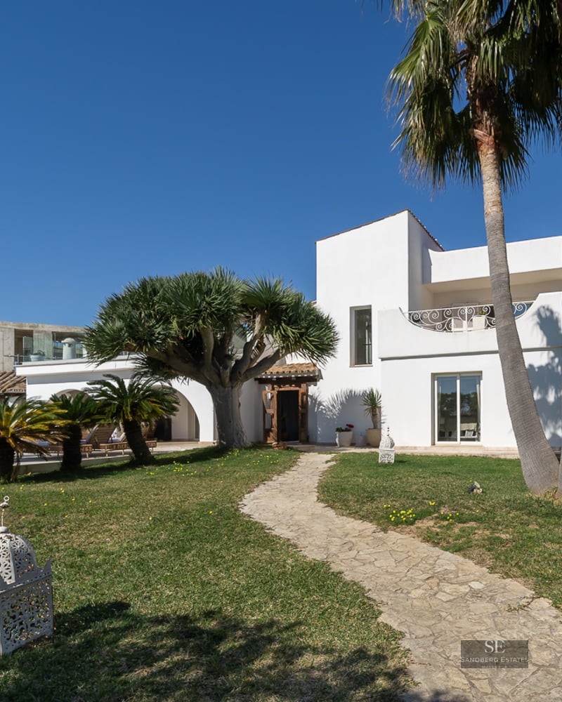 White Mediterranean villa with a winding stone path, palm trees, and a swimming pool under a bright blue sky.