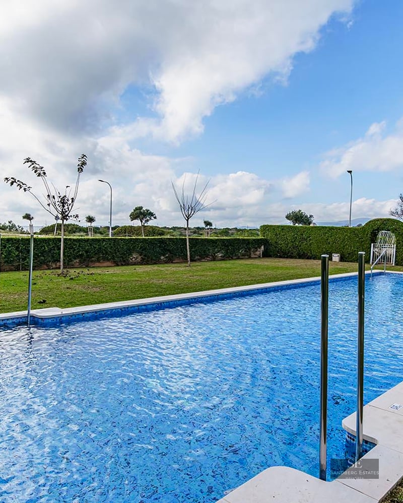Large rectangular blue-tiled swimming pool surrounded by a green lawn and manicured hedges under a cloudy sky.