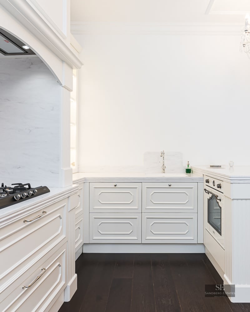 Elegant white kitchen with marble countertops, dark wood flooring, and a crystal chandelier.