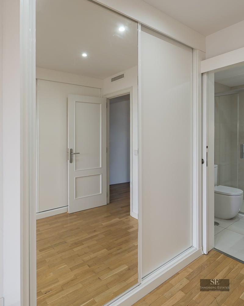 A bright modern room with light wood floors, a large mirrored sliding wardrobe, and a doorway leading into a white tiled bathroom.
