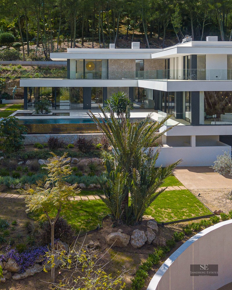 Aerial view of a three-story modern white villa with an infinity pool, landscaped gardens, and natural stone walls.