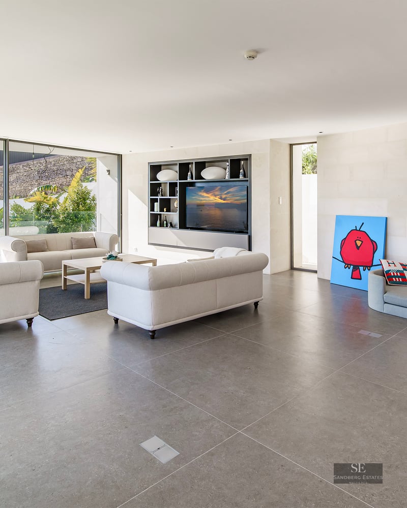 Bright modern living room with beige sofas, grey stone floor, and large glass windows overlooking a garden.
