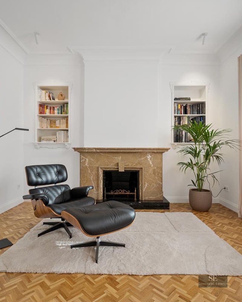 Modern living room with leather Eames chair, marble fireplace, parquet floor, and built-in bookshelves.