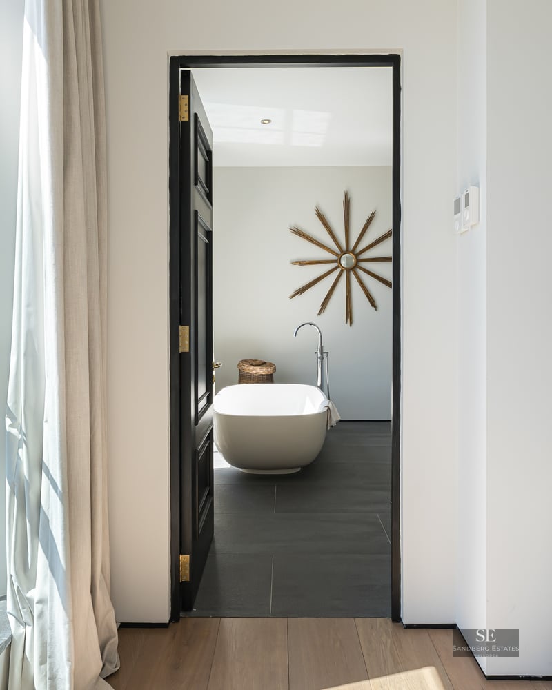 Open doorway revealing a modern bathroom with a freestanding tub and sunburst mirror, next to a window with an ocean view.