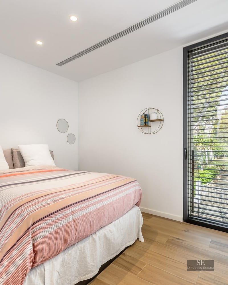 Bright bedroom featuring a striped bed, light wood flooring, and a large window looking out onto lush greenery.