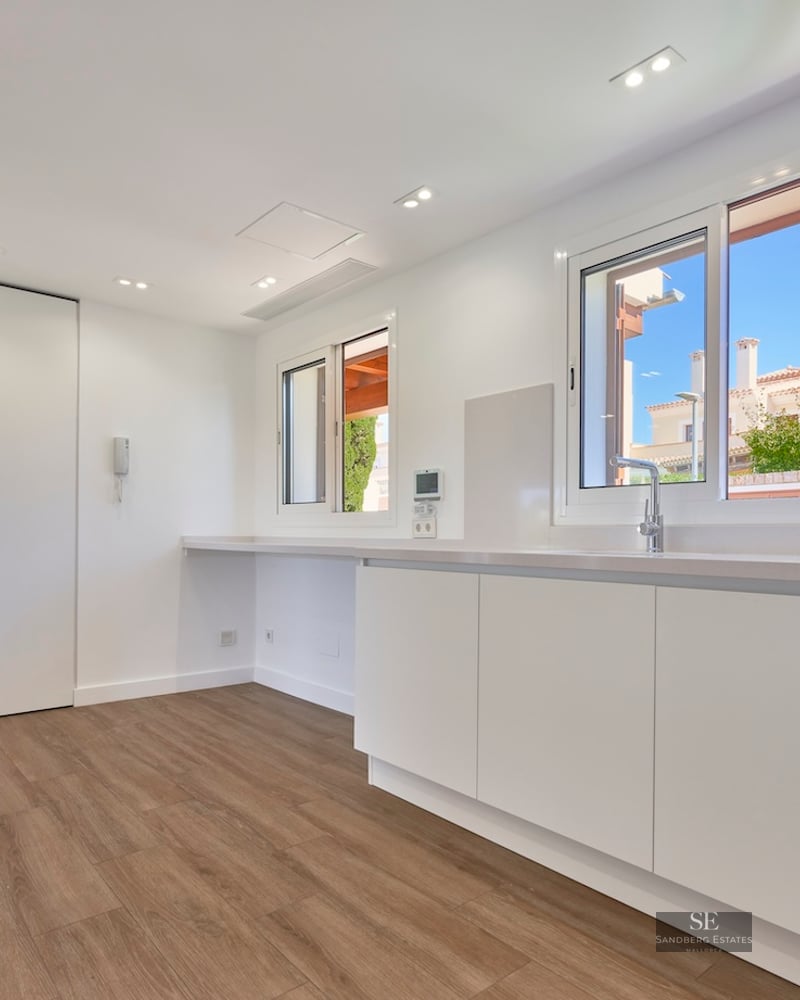 Modern kitchen with white cabinets, wood flooring, stainless steel sink, and bright windows.