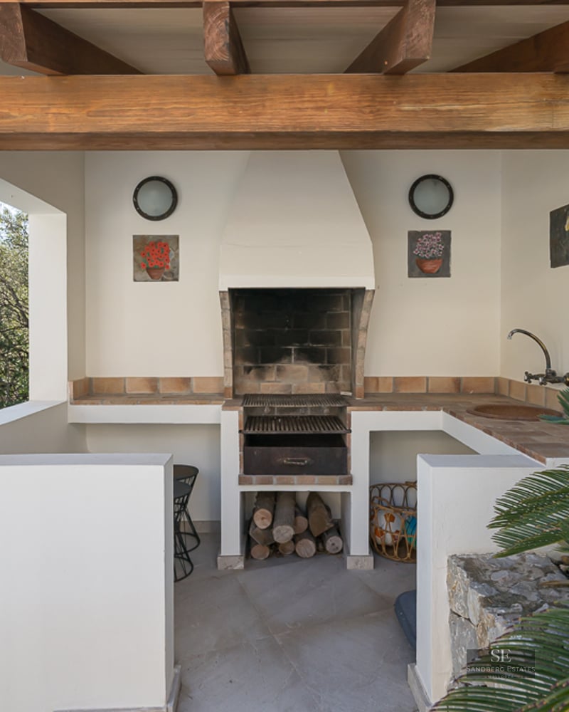 Un espace barbecue extérieur couvert avec des poutres en bois rustiques, des murs blancs, un gril en pierre et un évier.