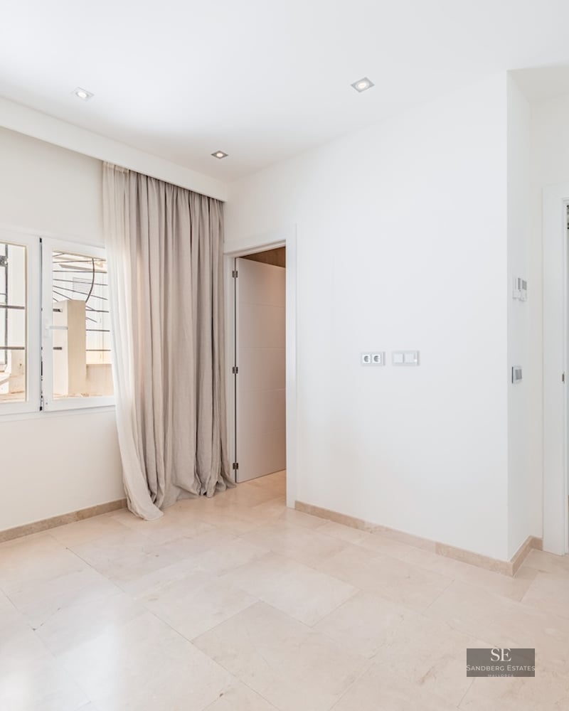 Bright empty room with white walls, cream marble floor, window with floor-length beige curtains, and open doorways.