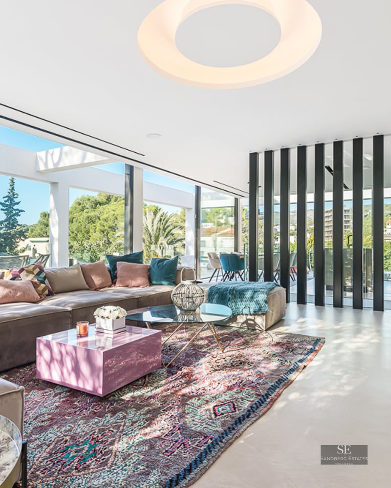 Modern living room featuring a large taupe sofa, colorful patterned rug, and floor-to-ceiling windows with greenery views.