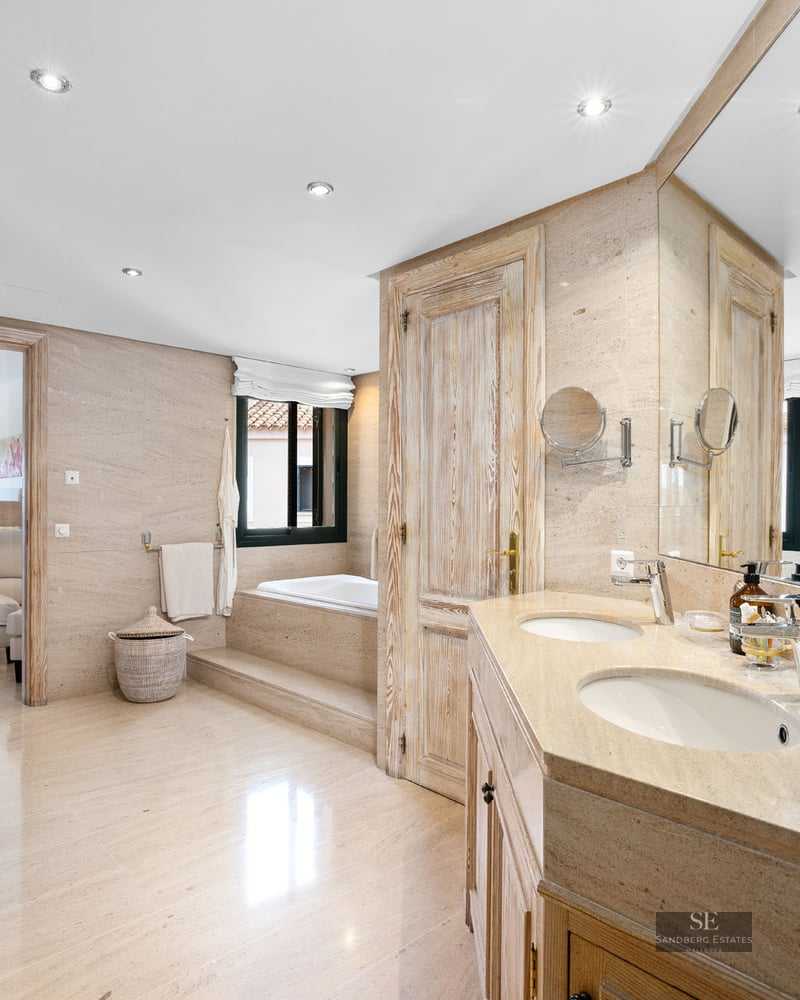 Salle de bain spacieuse avec murs en pierre beige, double vasque, grand miroir et baignoire encastrée au fond.