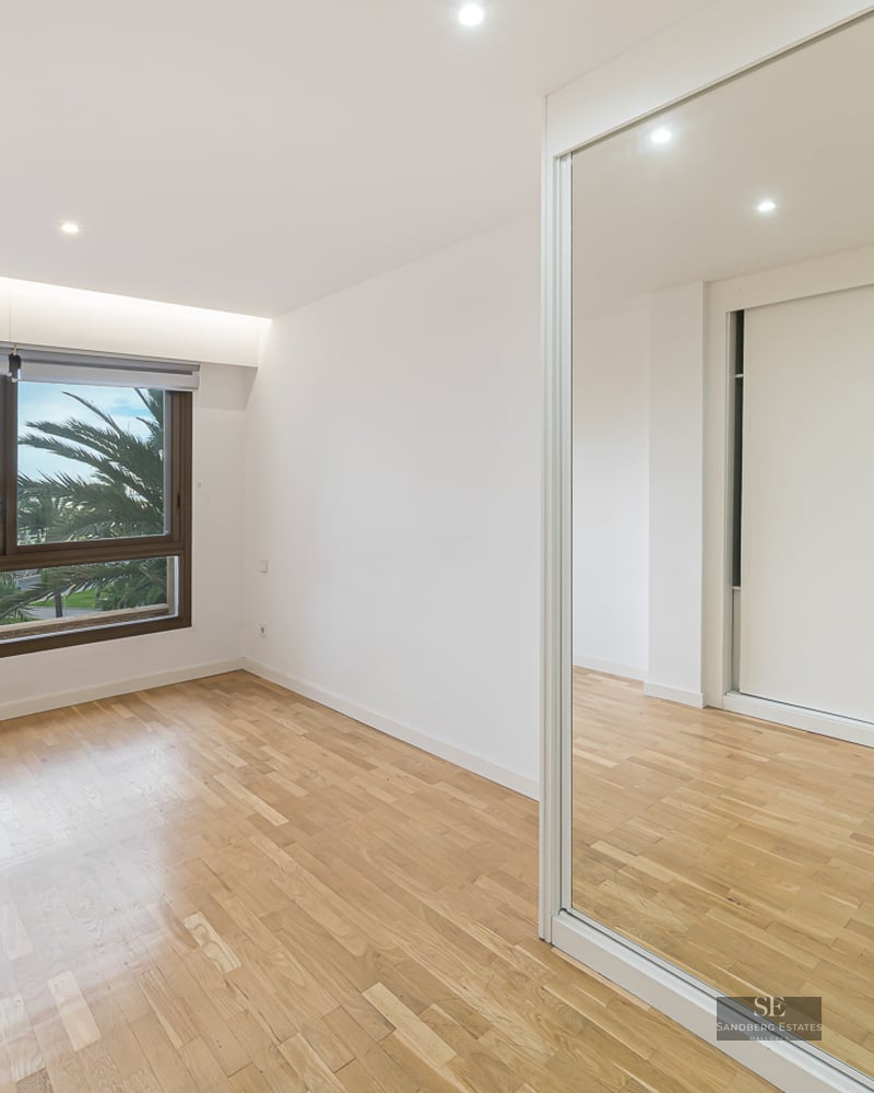Empty bedroom with light wood floors, mirrored wardrobe, and a window showing palm trees and boats.