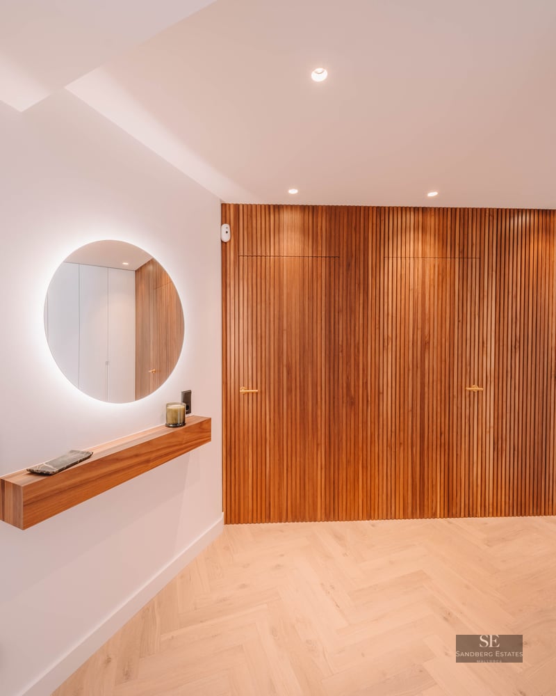 Minimalist hallway featuring a backlit circular mirror, wooden floating shelf, and vertical wood slat wall.