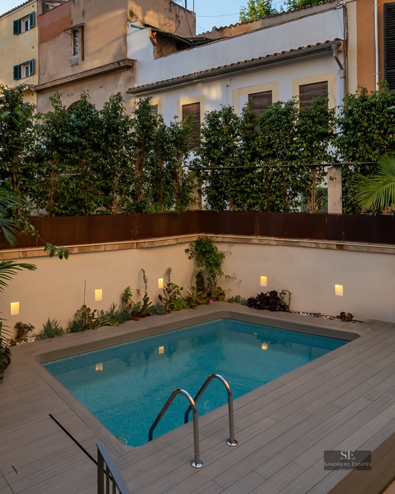 Modern rectangular pool with grey decking, surrounded by white walls with integrated lighting and lush greenery.