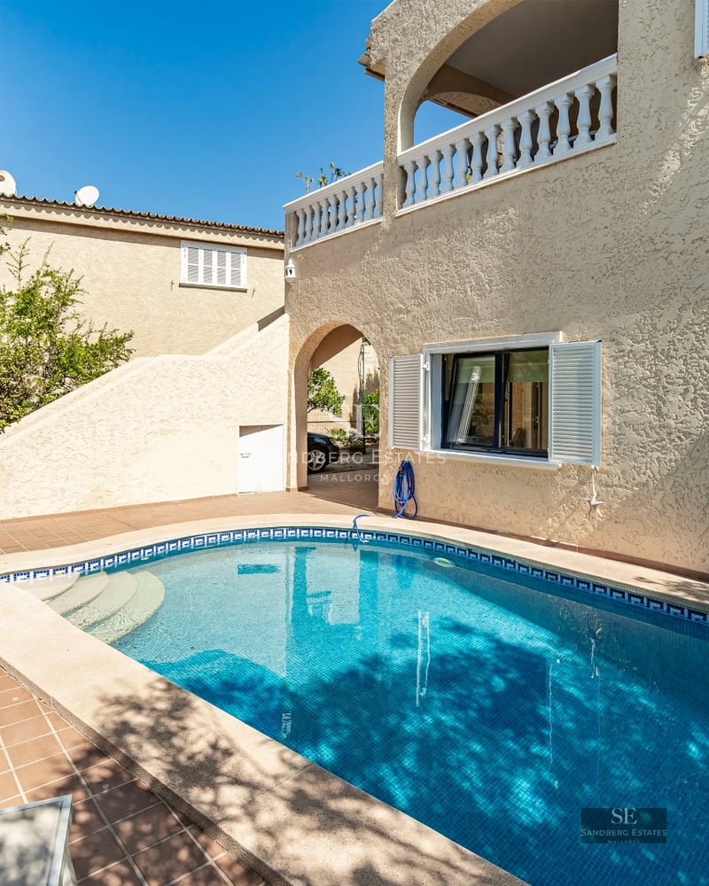 View of a pool surrounded by lawn and a modern two-story house with large windows. Natural lighting and minimalist design.
