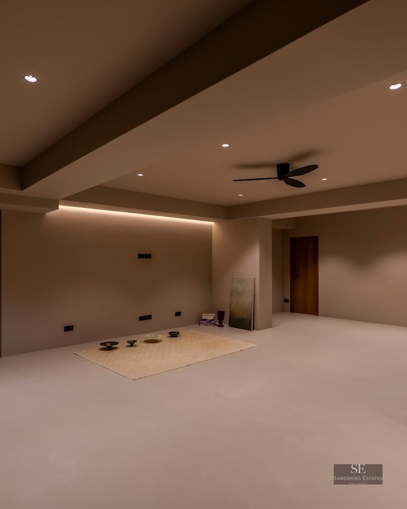 Une grande pièce minimaliste aux murs et sols beiges, avec éclairage encastré et ventilateur de plafond.