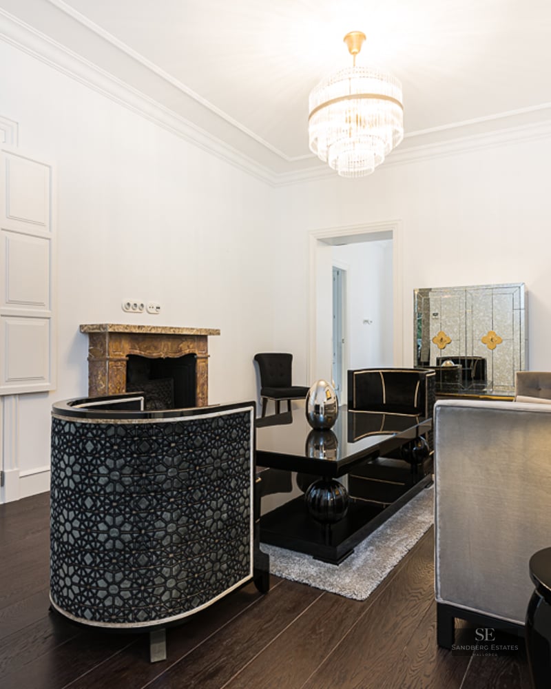 Elegant living room with white walls, dark wood floors, designer furniture, crystal chandelier, and a marble fireplace.