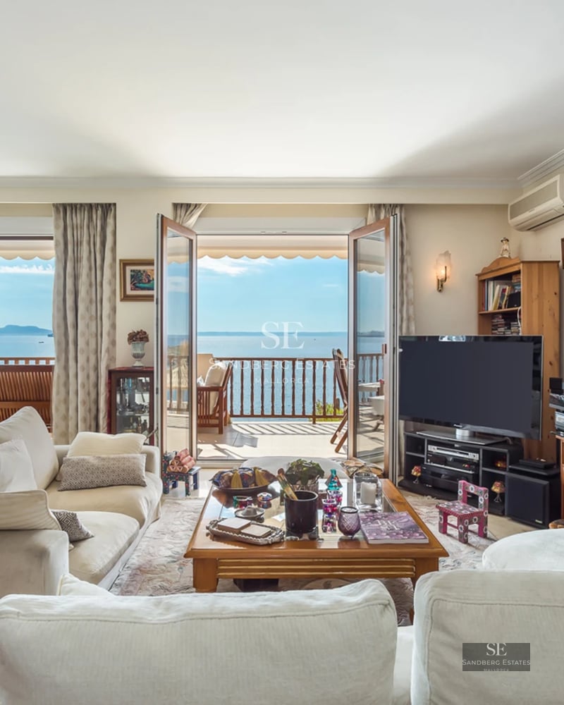 Bright living room with white sofas, wooden furniture, and glass doors opening to a balcony with an ocean view.