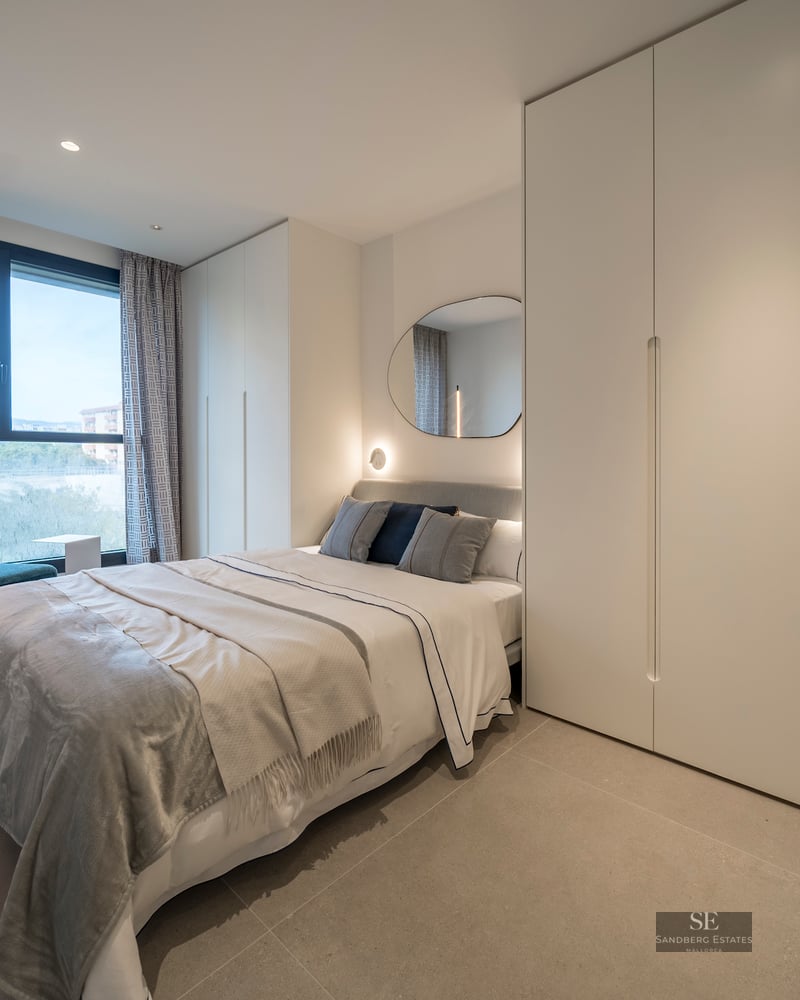 A bright modern bedroom featuring a bed with grey linens, large white wardrobes, a teal chair, and a large window.