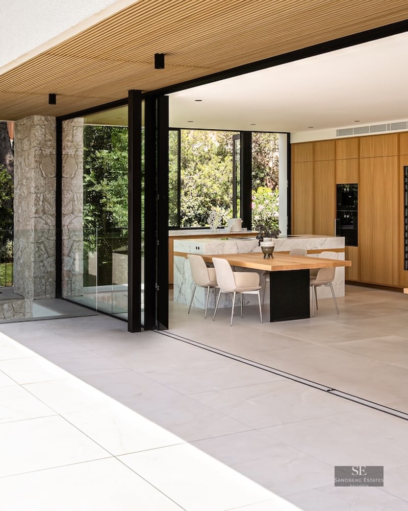 Modern kitchen with wood cabinets and marble island opening to a sunny stone terrace via large sliding glass doors.