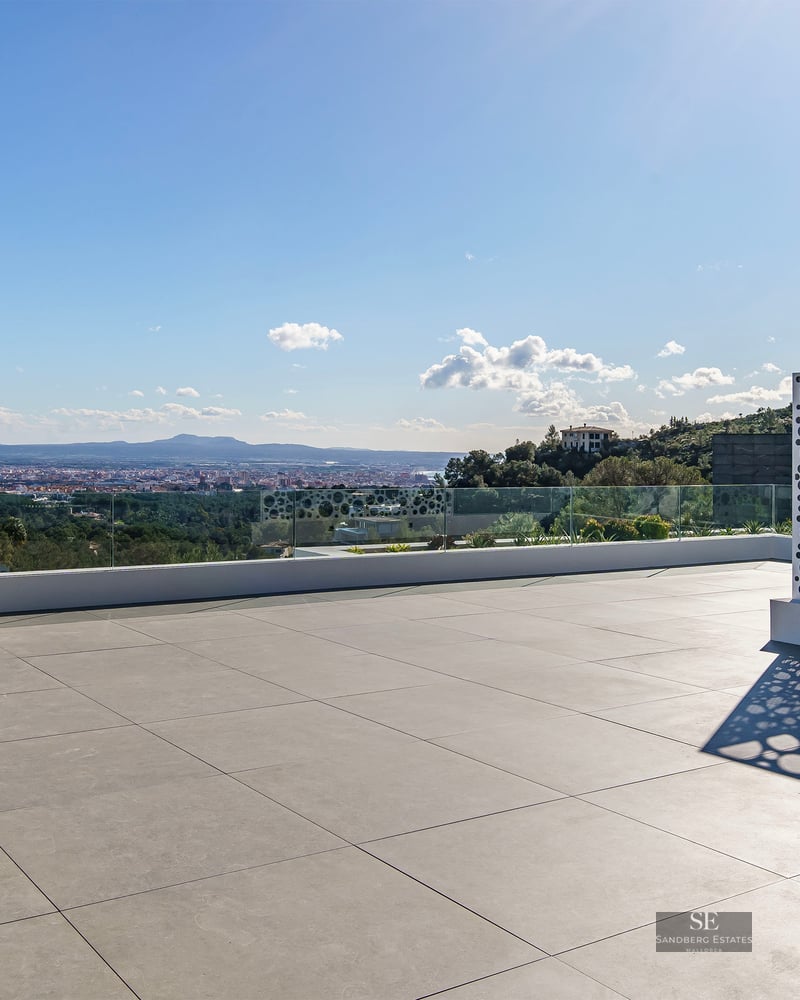 A spacious modern rooftop terrace with grey tiles, glass railings, and a white perforated screen overlooking a city.