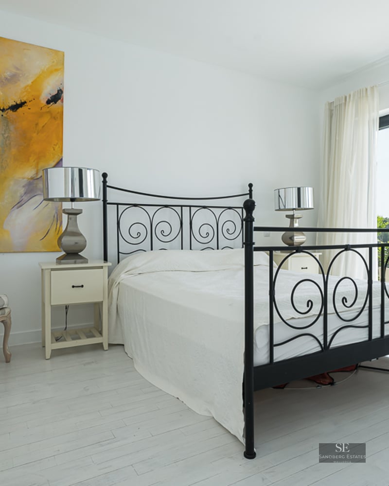 Bright white bedroom featuring a black wrought iron bed, abstract art, and large glass doors to a sunny terrace.