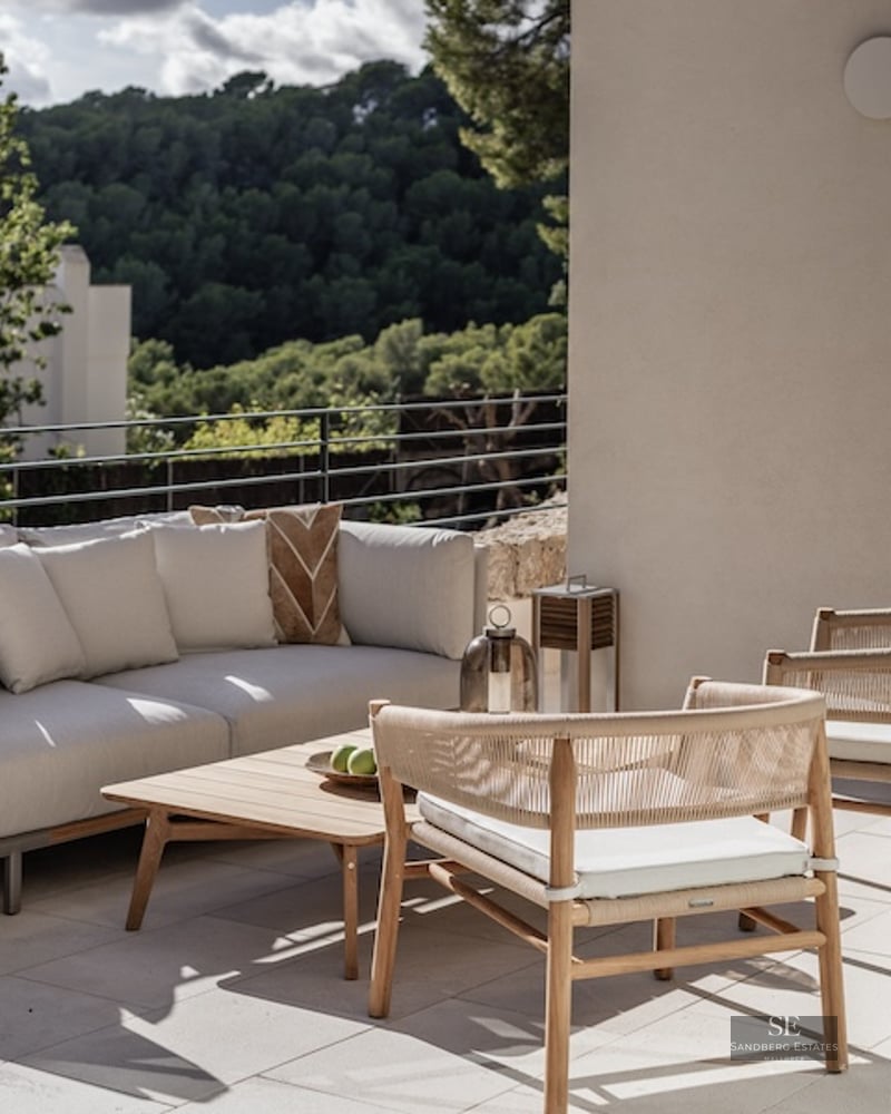 Modern outdoor seating area on a terrace with a white sofa, wooden chairs, and views of a lush green forest.