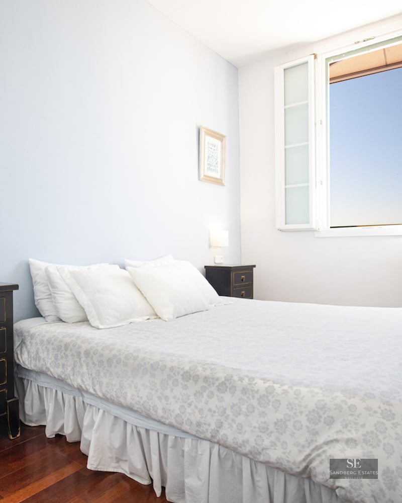 A bedroom with a white floral bed, black nightstands, dark wood floors, and an open window showing a blue sky.