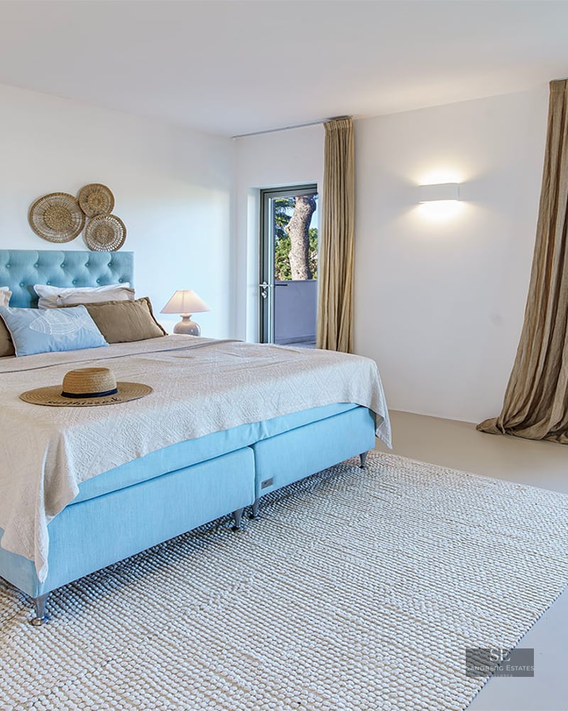 Bright room with light blue bed, wool rug, and large window overlooking green trees.
